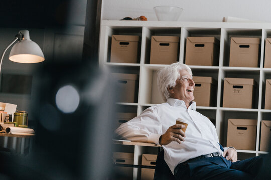 Laughing senior businessman with coffee to go sitting in his office