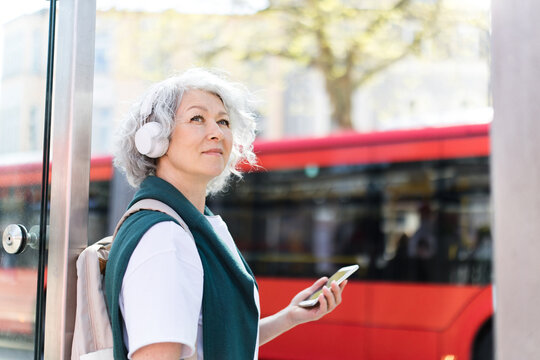 Woman with headphone and smartphone waiting at bus stop in city