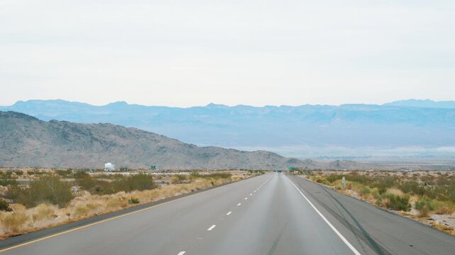 Pov driving on long straight asphalt road through an arid desert landscape with distant hazy mountains on horizon, embodying concept of classic american road trip. Transportation on interstate highway
