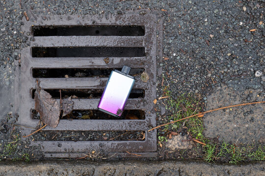A vibrant purple vape electronic cigarette is left lying on a wet metal drain cover in the rain.
