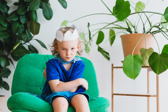 A grumpy boy wearing a paper crown sits on a green armchair indoors