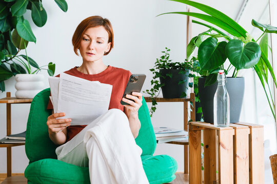 Focused woman reads documents and uses smartphone in her home office