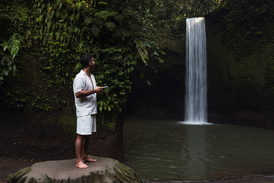 A male tourist holds a smartphone near a waterfall in Bali
