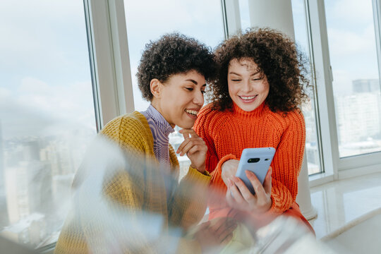 Two friends with curly hair smiling and looking at phone on windowsill
