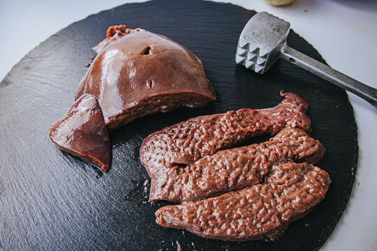 Raw liver pieces arranged on a dark cutting board beside a metal meat tenderizer, showing glossy surfaces and varied organ-meat textures.