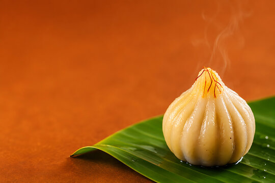 Single steaming modak sweet with saffron strands on a green banana leaf against a warm orange background, for ganesh chaturthi festival