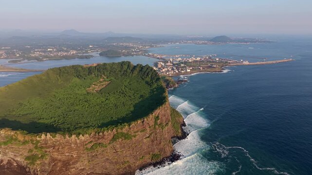 Aerial view of Seongsan Ilchulbong tuff cone with lush green crater and steep rocky cliffs overlooking the blue ocean in Seogwipo-si, Jeju-do, South Korea.