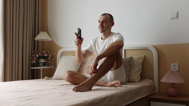 Young man sitting on bed, holds remote control, watching TV and smiling.