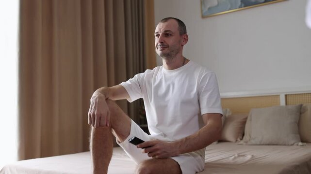 Young man sitting on bed, holds remote control, watching TV and smiling.