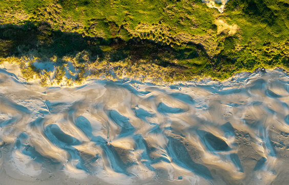 Aerial view of the sandy beach and dunes featuring intricate patterns in the sand, golden hour light, and lush green coastal vegetation in Bandon, Oregon Coast, United States.