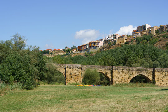 Scenic view of a rustic stone bridge spanning a green field beneath a sunny sky, with traditional buildings clustered on the sloping hill