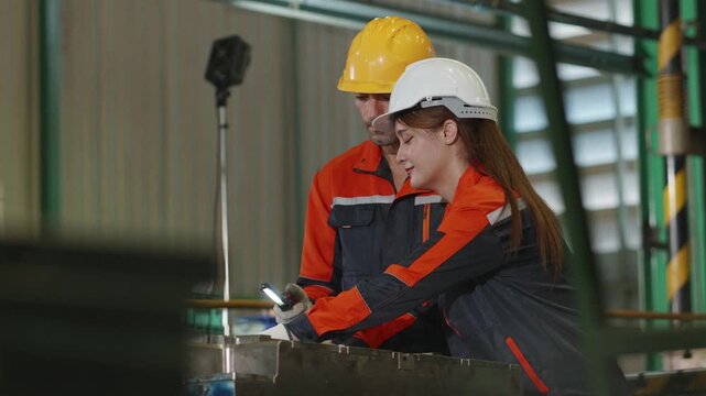 Two engineers in hard hats inspect industrial machine mold parts using a flashlight inside a factory workshop, checking metal equipment during quality control maintenance.