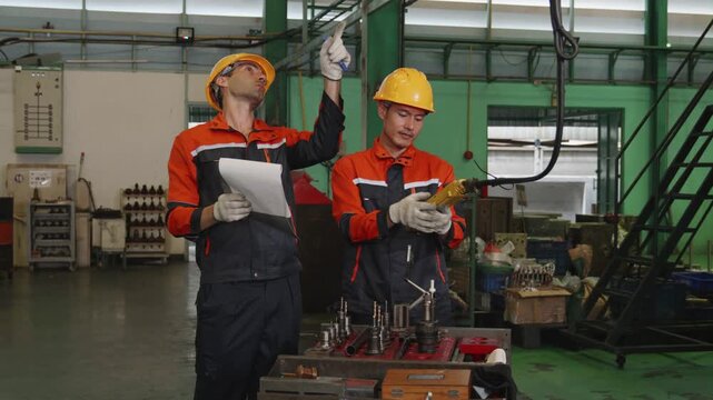 Two male engineers in yellow hard hats review a checklist and inspect precision tooling parts on a workbench, using a hand tool inside an industrial factory warehouse.