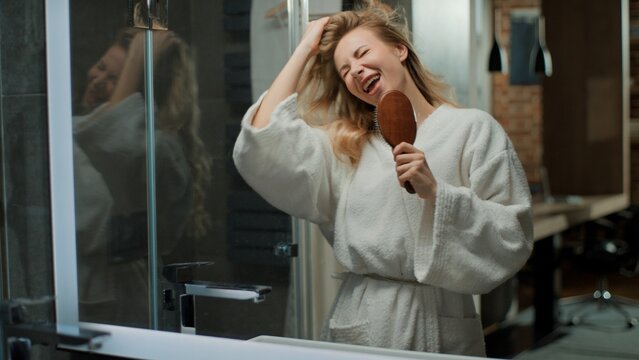 Funny happy Caucasian middle-aged woman singing in hairbrush touching soft shiny curly hair having fun fooling around looking at mirror reflection at home bathroom positive female sing in brush comb