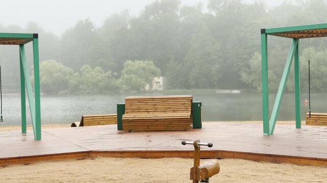 Wooden benches and pavilions stand on a misty lake shore in Saint Petersburg. Fog hangs over the quiet trees across the water. The serene playground creates a calm, peaceful mood.