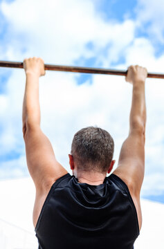 Athletic mature man doing pull ups on a horizontal bar outdoors from behind. Fit middle aged male training in a public park against a beautiful blue sky with clouds.