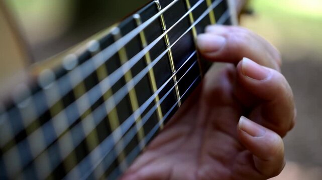 Hand changes chord positions along guitar fretboard in tight close-up, pressing metal strings against dark frets while shallow focus isolates precise finger movement outdoors