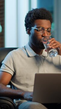 Concentrated healthy African American business man drinking water working laptop at night evening at home office ethnic male focused guy businessman worker drink refreshing work deadline with computer