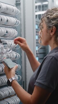 Female Engineer Audits Rack Connections Using Smartphone, Comparing Port Labels With Digital Records While Male Colleague Services Nearby Equipment During Infrastructure Verification. Vertical Shot.