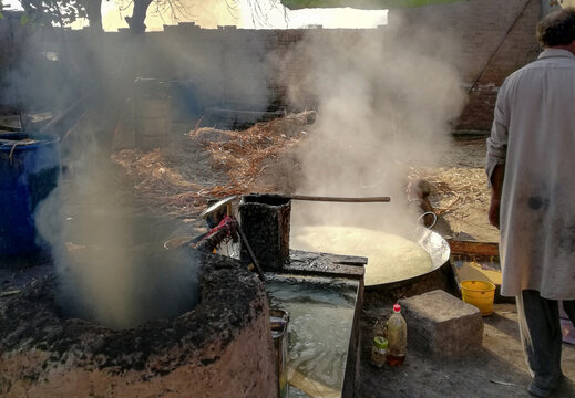 View of a man overseeing the traditional production of jaggery in a large boiling vat with rising steam in a rural workshop in Multan, Punjab, Pakistan.