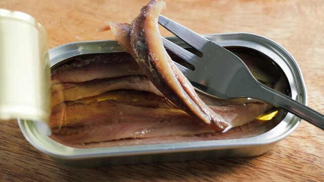 Canned anchovy fillets in oil inside an open metal tin with a fork lifting one fillet on a wooden table. High quality 4k footage
