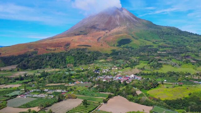 Aerial view of Mount Sinabung volcano towering over the village of Naman Teran with lush green agricultural fields and surrounding tropical landscape in Naman Teran, North Sumatra, Indonesia.