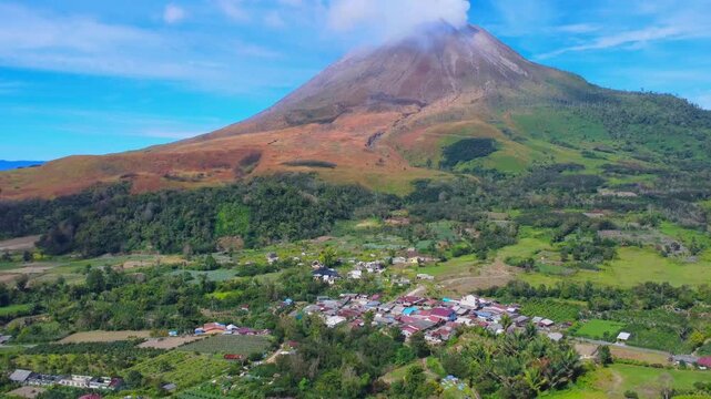 Aerial view of Mount Sinabung rising above the village of Naman Teran, surrounded by lush green agricultural fields and volcanic slopes in Naman Teran, North Sumatra, Indonesia.