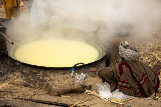 View of a man in traditional attire sitting by a large vat of boiling sugarcane juice during the traditional jaggery production process in Multan, Punjab, Pakistan.