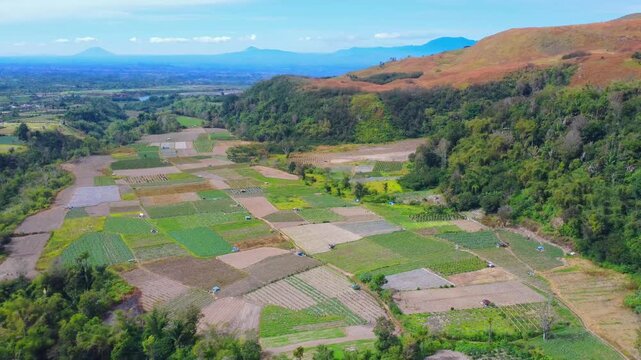 Aerial view of Mount Sinabung overlooking vibrant patchwork agricultural fields and lush hills in Naman Teran, North Sumatra, Indonesia.