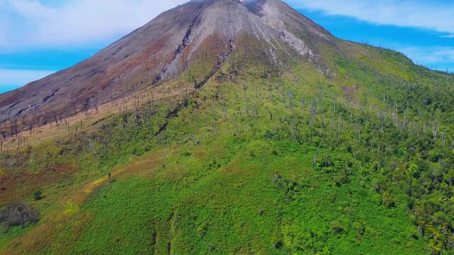 Aerial view of Mount Sinabung with its volcanic slopes, lush green vegetation, and rocky terrain under a bright blue sky in Naman Teran, North Sumatra, Indonesia.