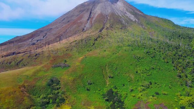 Aerial view of Mount Sinabung with lush green slopes and rocky volcanic summit under a clear blue sky in Naman Teran, North Sumatra, Indonesia.