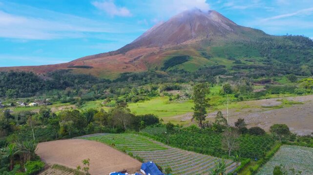 Aerial view of Mount Sinabung volcano rising above lush green agricultural fields and a small house in Naman Teran, North Sumatra, Indonesia.
