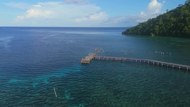 Aerial view of a long wooden pier extending into the turquoise waters of Raja Ampat Regency, surrounded by lush green islands, Kri Island, Southwest Papua, Indonesia.