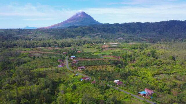 Aerial view of Mount Sinabung volcano rising above the lush green fields, winding roads, and scattered houses in Naman Teran, North Sumatra, Indonesia.