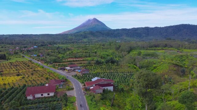 Aerial view of Mount Sinabung volcano rising above the lush green landscape and rural road in Naman Teran, North Sumatra, Indonesia.