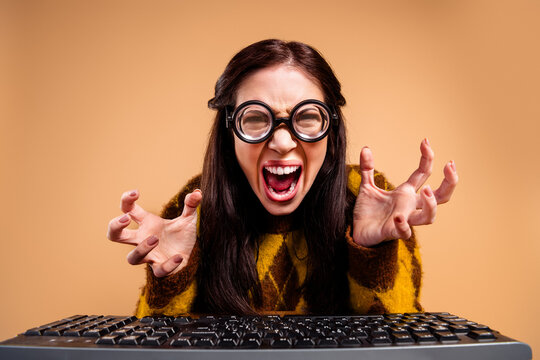 Frustrated young woman yelling at computer keyboard while wearing round glasses and a cozy sweater, expressing anger, stress and a dramatic tech meltdown in a studio portrait
