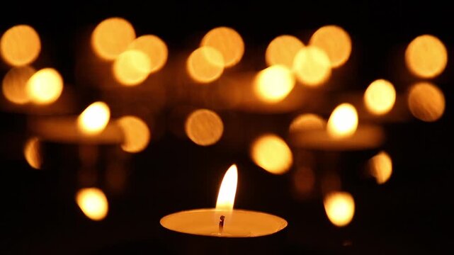 A lit candle burns steadily in sharp focus at the bottom of the frame while rows of golden bokeh lights flicker softly above against a dark blurred background throughout.