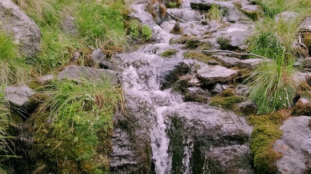 Mountain stream flows over gray rocks through a green valley in the Fagaras Mountains near Transfagarasan in Romania beneath white clouds and blue sky