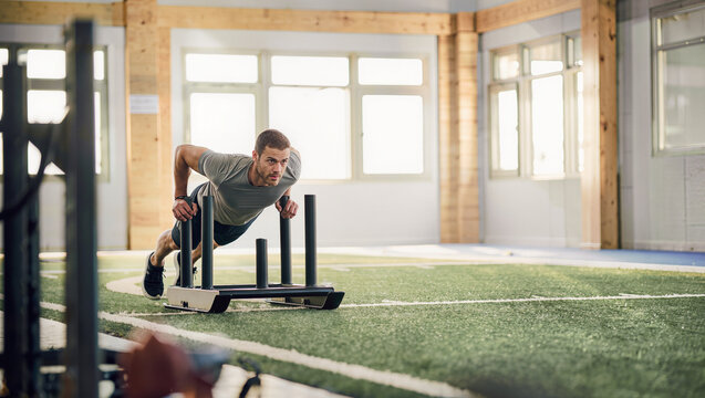 Fit man pushing a weighted sled across indoor turf during a gym workout, functional strength training and athletic fitness motivation with copy space