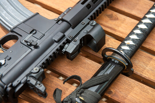 A modern semi-automatic rifle with a red dot sight lying alongside a traditional Japanese katana sword on a wooden slatted table.