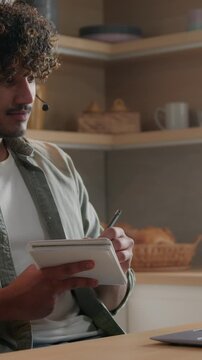Focused Indian Arabian business man wearing headset teaching online at home laptop video call conference writing notes remote lesson ethnic male tutor education student learning internet class indoors