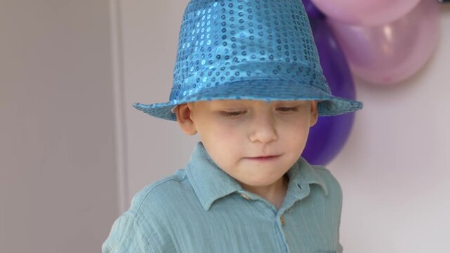 Boy with blue party hat smiles and lowers blowout after birthday celebration, showing happy childhood milestone beside pastel balloons and playful party decor, wearing sequined costume hat.