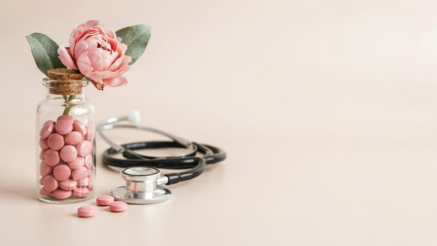 Holistic medicine concept with pink pills in glass bottle and stethoscope. Natural healthcare supplements and floral wellness treatment on beige background.