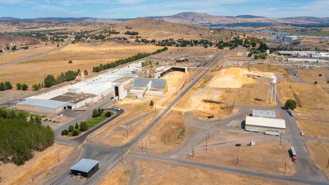 Aerial view of the Collins Products LLC industrial facility featuring large warehouses, wood chip piles, and water towers in the dry landscape of Klamath Falls, Oregon, United States.