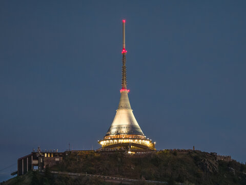 Aerial view of the iconic Jested Tower illuminated at night, perched atop the mountain peak against a dark blue twilight sky in Liberec District, Liberec Region, Czechia.