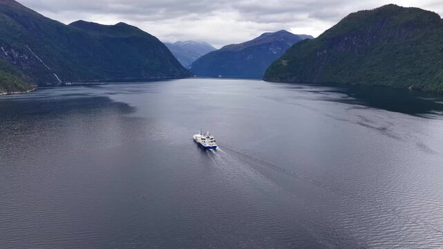 Aerial view of a wellboat navigating through the calm waters of a deep fjord surrounded by steep mountains under a cloudy sky in Liabygda, More og Romsdal, Norway.