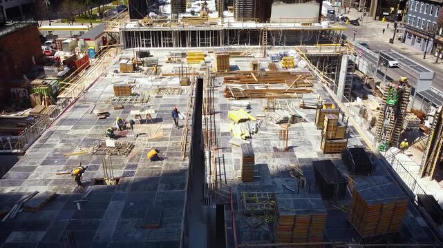 Overhead view of urban construction site with workers walking across concrete slabs and arranging stacked plywood formwork for mid-rise building columns