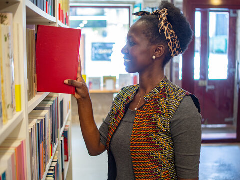A smiling African woman placing a red hardcover book back onto a white bookshelf in a modern library or bookstore.