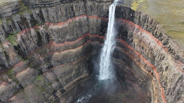Aerial view of Hengifoss waterfall, showcasing its iconic red clay layers embedded in basaltic rock strata, with water cascading into the deep gorge in Iceland.