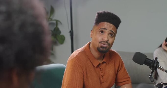 African American man in rust polo reacting to senior speaker, speaking into mic in podcast studio